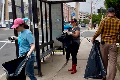 Three people in an urban setting near a sidewalk bus shelter use black bags to collect litter.