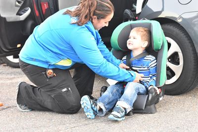 in an outdoor setting at the open door of  a car, a woman with a ponytail and blue top, black jeans, kneels on one knee while adjusting the straps on a carseat which is sitting on the pavement. In the carseat and smiling at the woman is a young boy in jeans and a blue, striped, long sleeve shirt. 