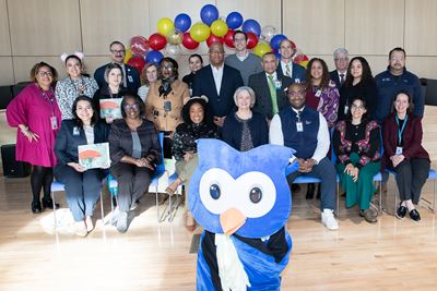A large group of people in business dress, seated and standing, pose in paneled room in front of blue, yellow  and speckled balloons. In front of them is the Reading Across America mascot, a cartoonish blue owl with big eyes.  