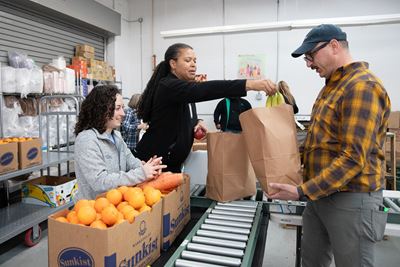 two women standing on one side of a conveyer belt, handing out fruit, whiel a man receives bananas in a paper bag on the other. Setting is a warehouse/garage with shelves of food items.