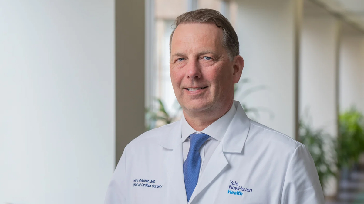A smiling, white man with greying brown hair in a white physician's jacket and wearing a blue tie. Background is a blurred hallway with windows and standing planters. His jacket is embroidered with the words Yale New Haven Health on left breast and Marc Peletier, MD, Chief of Cardiac Surgery on right.