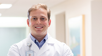 A smiling young white man with short blond hair poses in a hospital hallway. He is wearing a white clinical jacket  with an id badge.