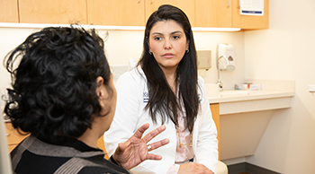 Two women, seated in a hospital exam room, face each other in discussion. In the foreground, one woman, in plaid, faces away and gestures with her left hand. The other, in a white clinical jacket, faces the camera. 