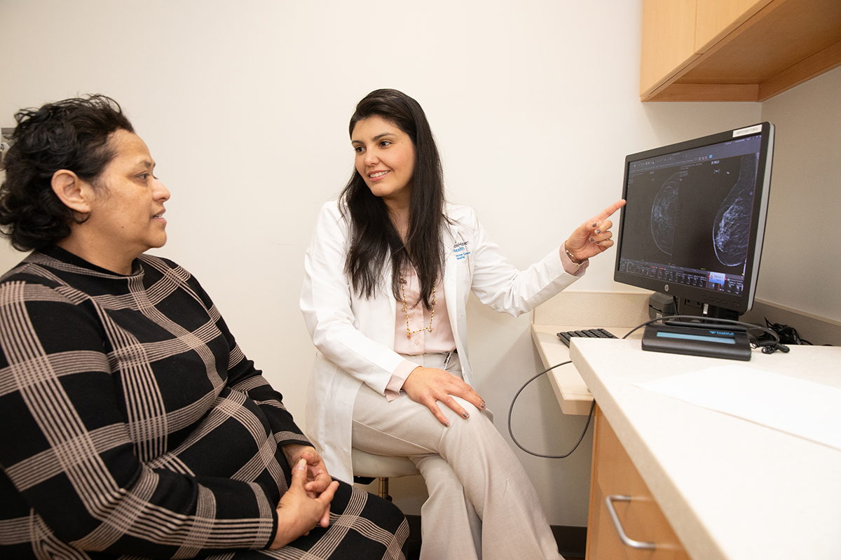 A seated, smiling woman a white jacket and tan slacks points to an image on a computer monitor. She faces another woman to her left, who is dressed in a plaid outfit, who is looking at the same screen.